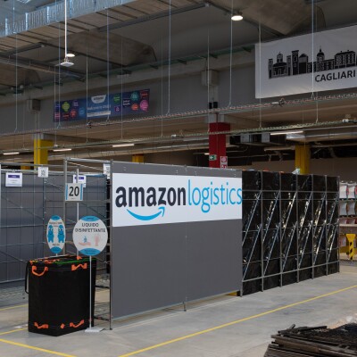 Foto di un'area di un magazzino Amazon. Su un pannello al centro si vede il logo di Amazon Logistics, mentre in fondo uno striscione con il profilo di una città e la scritta Cagliari