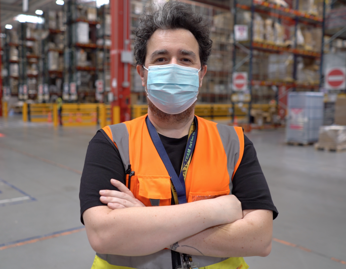 Uomo ripreso a mezzo busto in un magazzino Amazon. Indossa un giubbino catarinfrangente arancione e giallo, ha le braccia conserte. Indossa una mascherina chirurgica sul volto, ha i capelli neri con un ciuffo bianco. Alle sue spalle si intravedono gli scaffali di un magazzino Amazon.