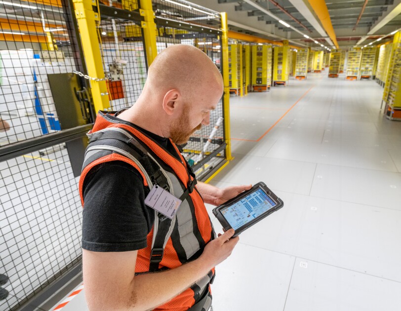 Ein Mitarbeiter mit Sicherheitsweste und Tablet steht in einem Bereich mit Transportrobotern.