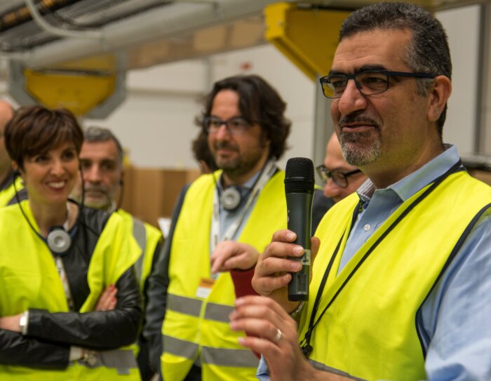 Gruppo di uomini e donne all'ingresso del Centro di Distribuzione Amazon a Piacenza. Indossano giubbini catarinfrangenti gialli. In primo piano, Tareq Rajjal, Amministratore Delegato di Amazon Italia Logistica, regge un microfono e parla ai presenti.