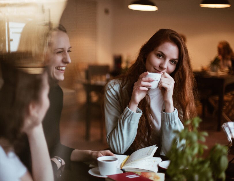 Eine Frau mit langen braunen Haaren sitzt an einem Tisch und lächelt in die Kamera. In ihren Händen hält sie eine Kaffeetasse.