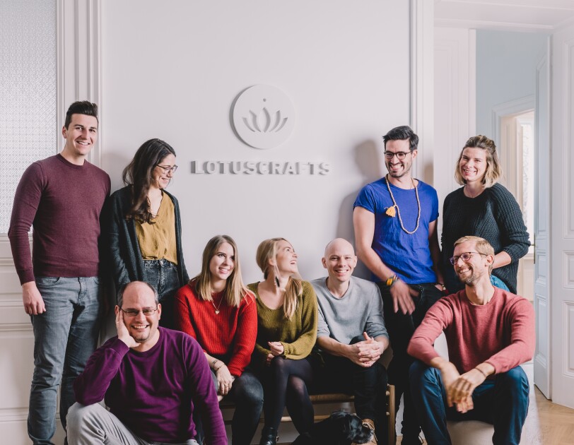 Das Team von Lotuscrafts sitzt und steht vor einer weißen Wand und lacht in die Kamera.