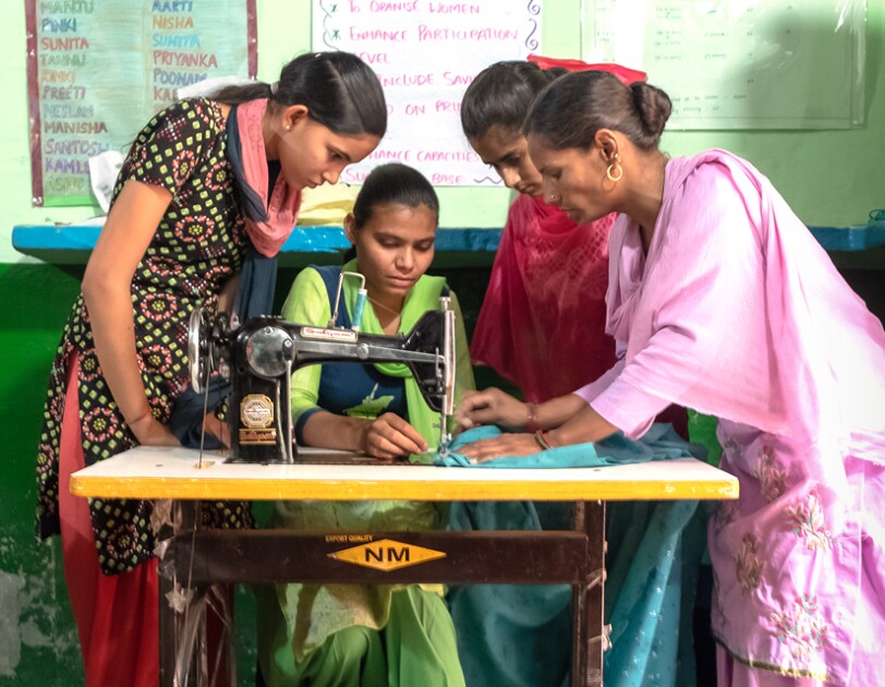 Three women stand around a woman who sits at a sewing machine.
