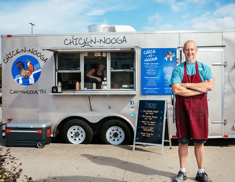 A man wearing a maroon apron stands with his arms crossed, in front of a silver CHICK-N-NOOGA food truck. A sign displays the specials in neon orange text on a black background.