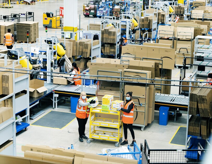 Wide shot of Amazon fulfilment centre staff stacking a yellow cart with medical supplies in the Coalville Fulfilment centre to provide supplies to the relevant government centres. They are wearing protective gloves and face masks while maintaining a social distance.