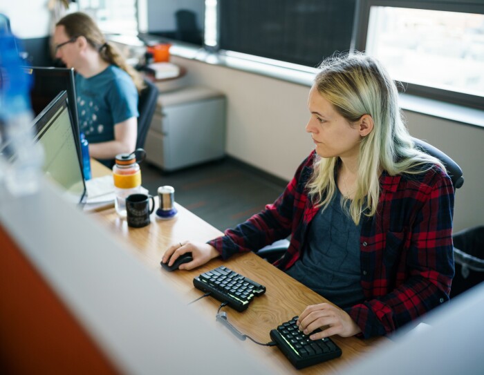 Danielle Skysdottir sits at her desk in a cubicle at Amazon's Seattle headquarters. She has blond hair and is wearing a T-shirt and flannel shirt. She's using a computer mouse with her right hand and typing on a split ergonomic keyboard with her left hand.