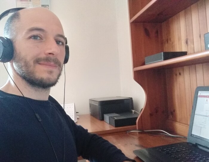 Primo piano di un uomo sulla sinistra. Indossa una maglia nera e delle cuffie, siede di fronte a una scrivania su cui è posato un PC.