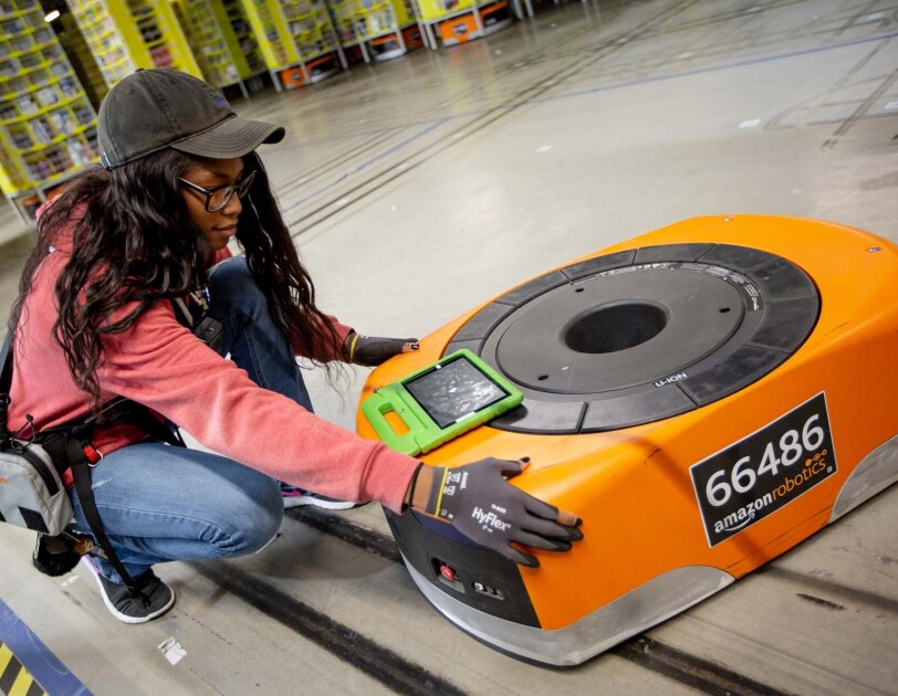 A woman kneels next to a robot, while also working on a tablet, at a Columbus, Ohio fulfillment center, CMH1