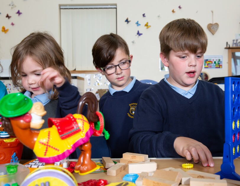 Three school children playing various games on a table, there is Connect Four, Buckaroo, Jenga and others.