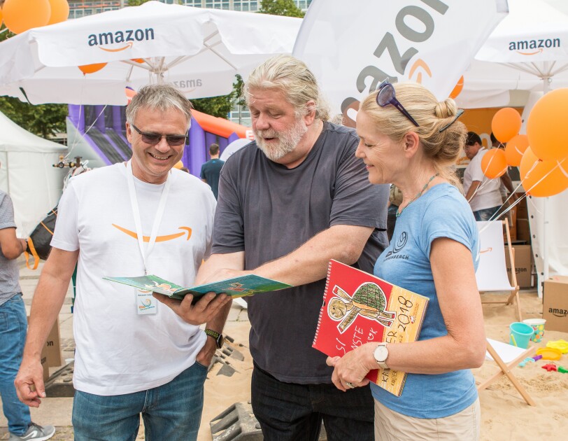 Rulo Lange ist in der Bildmitte zu sehen. Links Standortleiter Dietmar Jüngling, rechts eine Dame. Rulo Lange zeigt beiden ein KInderbuch. Im Hintergrund ist der SachsenBeach in Leipzig zu sehen, der von Amazon gesponsert wurde.