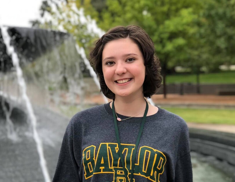 A young woman wearing a college sweatshirt stands in front of a fountain.