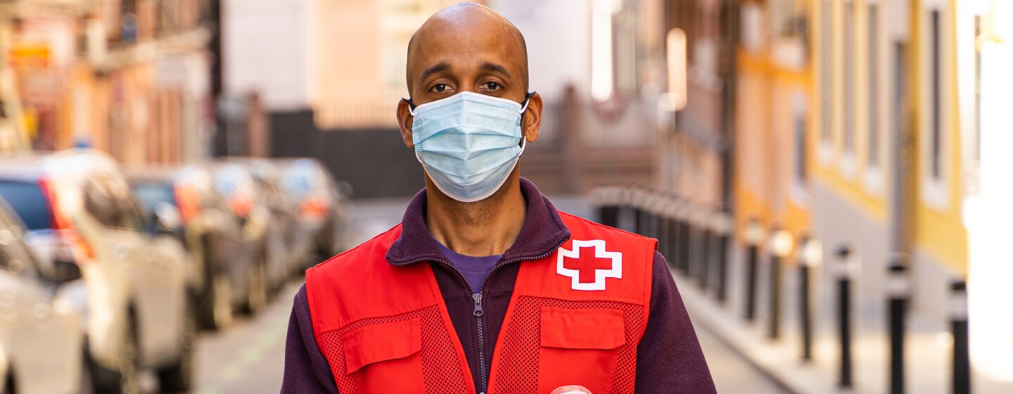 Alejandro Igor, voluntario de Cruz Roja. En una calle de Madrid. Él se encuentra en el inicio de la calle que hace cuesta. En un lado, desenfocados, coches aparcados y en el otro, también desenfocada, la acera. Él se encuentra en la mitad de la calle. Es moreno, lleva mascarilla azul y el pelo al cero. Vestimenta: con jeans de color azul, camiseta morada, polar morado y el chaleco de color rojo de la Cruz Roja. Mira a cámara y agarra con las dos manos dos cajas de cartón que són juguetes y encima también se puede apreciar algunos juguetes empaquetados en plástico,