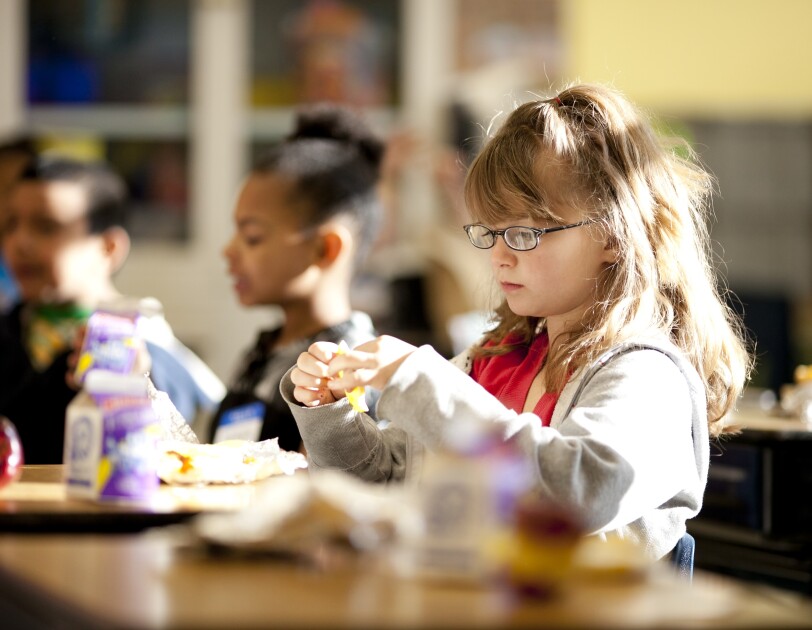 A young girl is opening preparing to eat. Behind her, other students are also eating. Apples and single-serve milk containers are on the desks in front of the students.