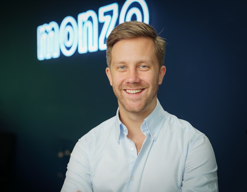 Tom Blomfield from Monzo looking directly at the camera and smiling against a blue background and Monzo logo