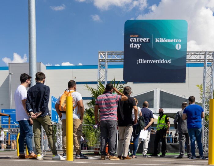 30 000 personas solicitaron un puesto de trabajo en Amazon durante el Career Day. En primer plano un grupo de 15 personas de espaldas a la cámara haciendo cola para participar en el Career Day de Amazon 2021. De fondo el centro logístico de Amazon en Illescas. La gente está haciendo cola, en la entrada, delante de un cartel que está apoyado en la parte superior de una columna, En el cartel pone ¡Bienvenido!, kilómetro 0 y Amazon Career Day. Se ve el cielo azul con nubes blancas.