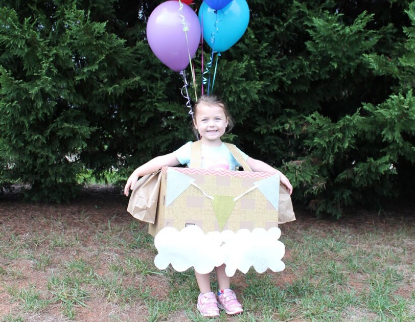 A young girl in a hot air balloon Halloween costume. She wears a box with pennants at the top, and paper clouds at the bottom. Above her float several colorful balloons.