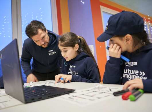 Ein Amazon Mitarbeiter erklärt Kindern  das Coden. Alle tragen Kapuzensweater mit mit Aufdruck Amazon Future Engineer.