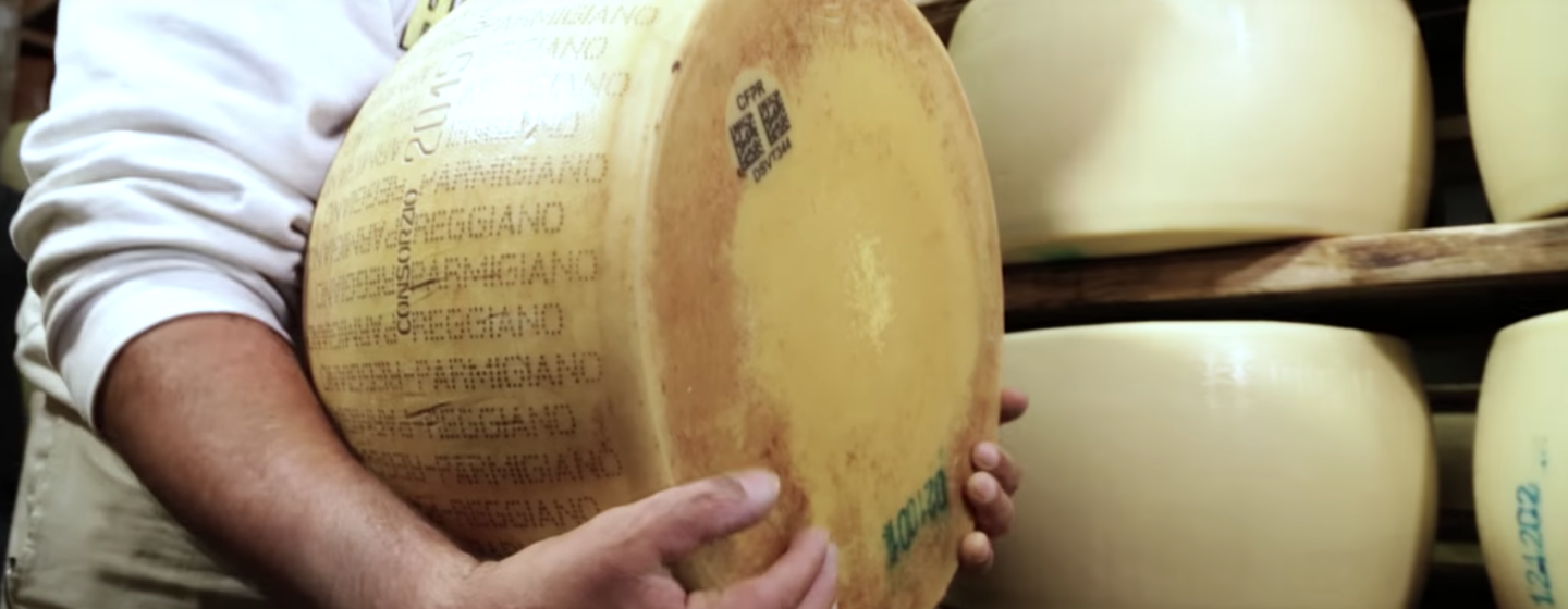 Primo piano di una forma di Parmiggiano Reggiano retta dalle mani di un uomo di cui non si vede il volto. Sullo sfondo, due scaffali con alcuni formaggi non stagionati.