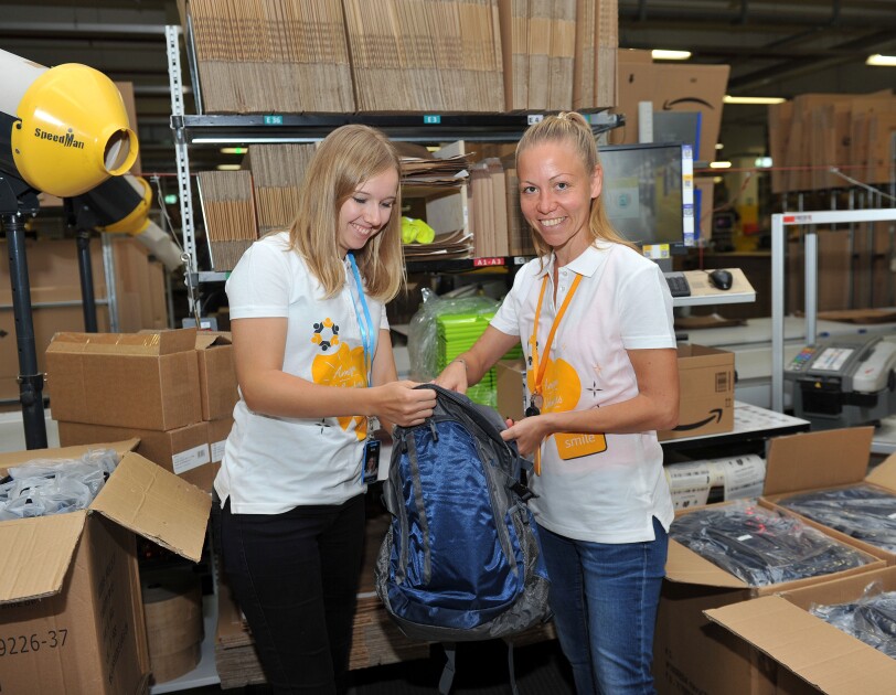 Zwei MItarbeiterinnen von Amazon, beide tragen ein weißes Poloshirt mit orangenen Herzaufdruck und der Beschriftung "Amazon Volunteer" stehen an einer Packstation, umgeben von Kartons. Sie halten in der Hand einen blauen Rucksack, den sie gerade beide mit verschiedenen Untensilien befüllen.