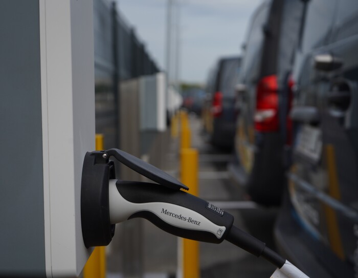 Una Mercedes Benz-Vans en un aparcamiento. Se ve el logo Prime de Amazon y el texto en la puerta 100% Eléctrico Envíos 'Shipment Zero'. En primer plano se ve un cargados de batería que está subministrando electricidad a la furgoneta de al lado que solo se ve un poco su parte trasera.