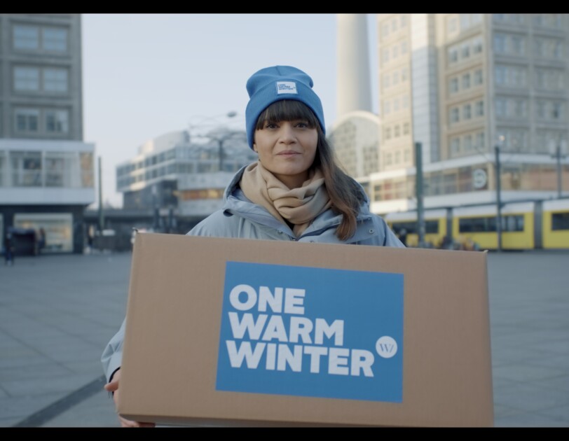 Eine dunkelhaarige Frau mit blauer Mütze schaut in die Kamera. Sie steht auf einem Platz und hat eine Winterjacke an. In der Hand hält sie einen Karton auf dem "One Warm Winter" steht.