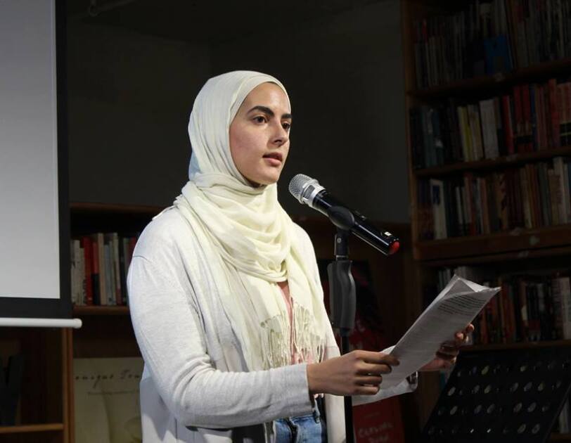 A woman (Sumaya Awad) stands in front of a microphone, speaking about her experience