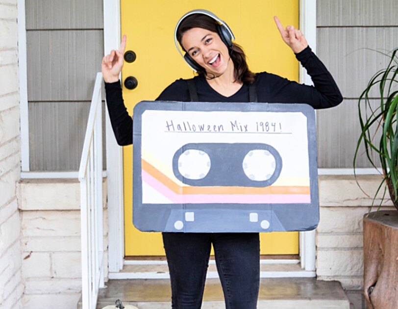 A woman wears jeans and a long-sleeve tee. over her torso is a box painted to look like a cassette tape, with "Halloween Mix 1984" on the front