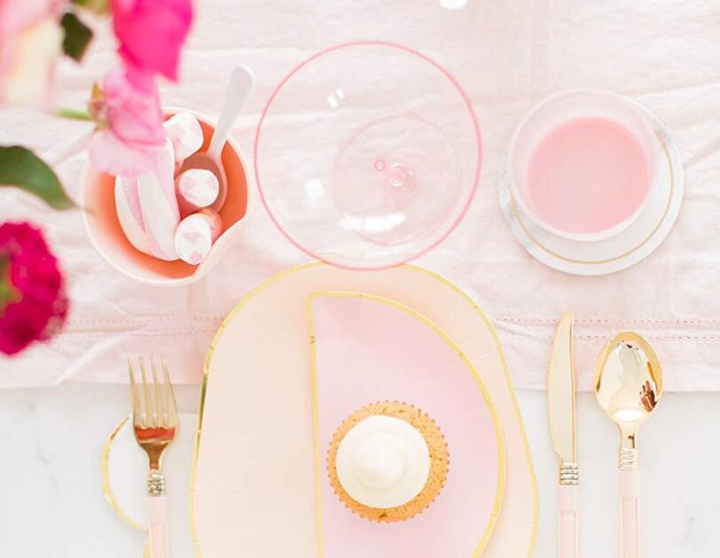 Pink champagne glasses sit atop a pink table runner on a marble table top. At each place setting, elegant dishes trimmed in gold and golden cutlery sit.