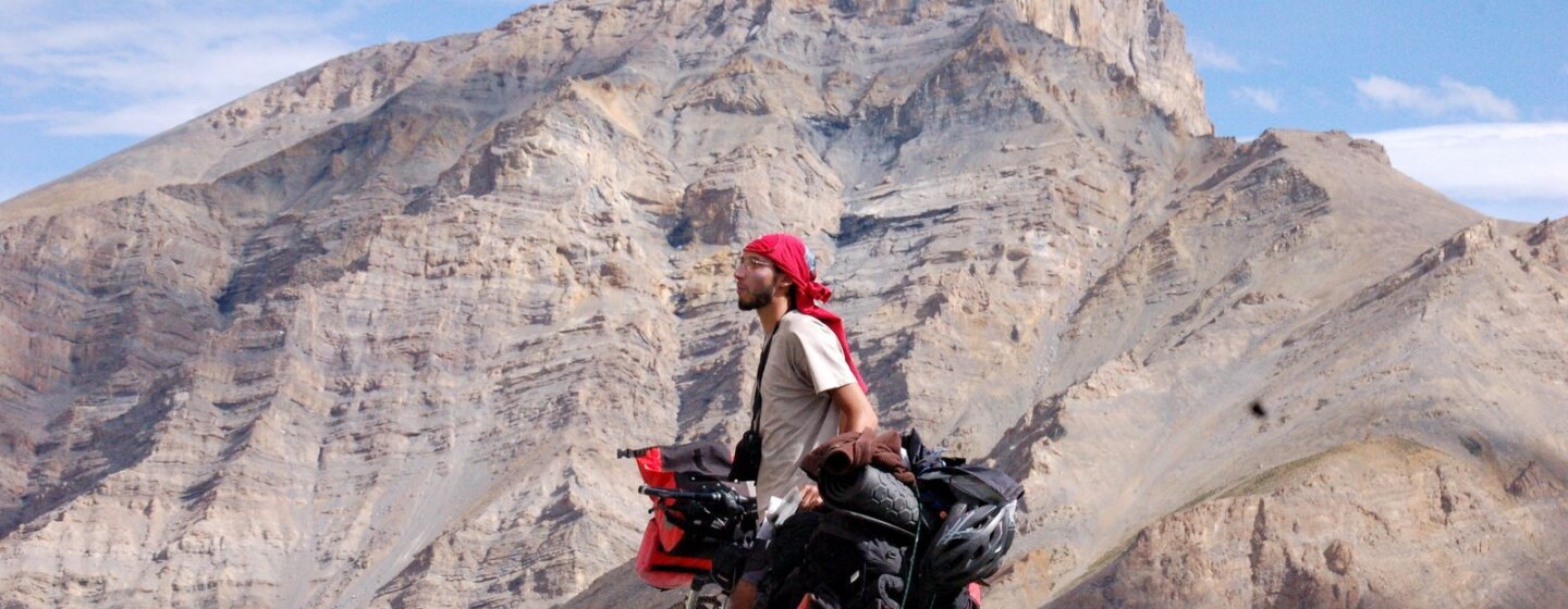 Karim Fahssis se tient à côté de son vélo chargé de sacs de voyage au milieu d'un paysage désertique. Il porte un foulard sur la tête. Derrière lui une montagne aride.