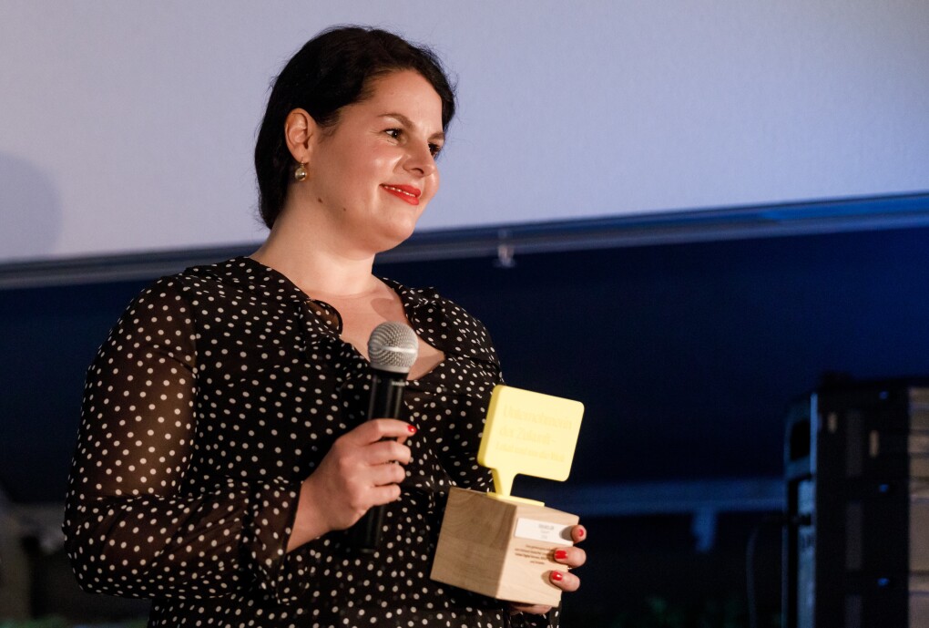 Julia Ritte hat den Preis steht auf der Bühne. In einer hand hält sie ein Mikrofon, in der anderen den UdZ-Pokal. Sie blickt freundig und gerührt ins Publikum.