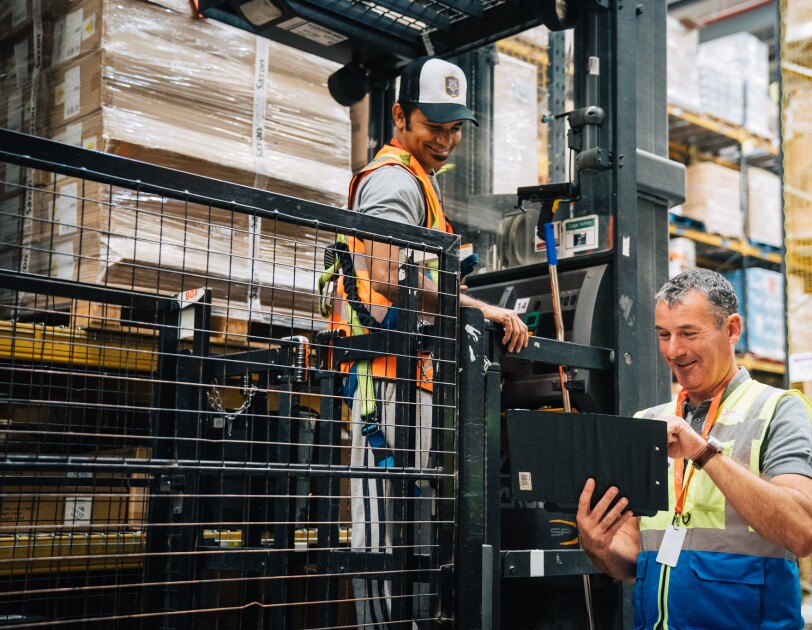 Temporary associate John Sheerin smiling at a colleague in an Amazon Fulfilment Centre and typing on a tablet.