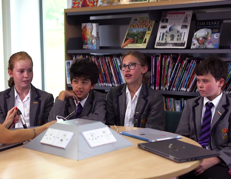 Students sitting around a table at school in front of a bookshelf, being interviewed by a BBC presenter