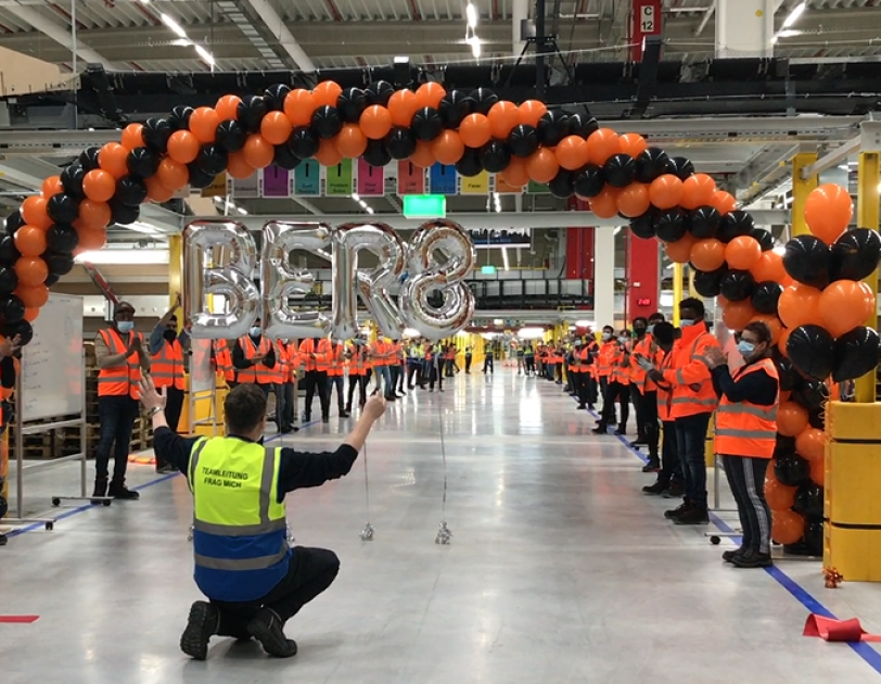 Mehrer Mitarbeiter mit Masken in einem Logistikzentrum: Alle klatschen, 2 Mitarbeiter halten einen Bogen mit schwarzen und orangenen Luftballons in die Höhe.