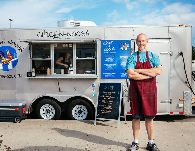 A man wearing a maroon apron stands with his arms crossed, in front of a silver CHICK-N-NOOGA food truck. A sign displays the specials in neon orange text on a black background.