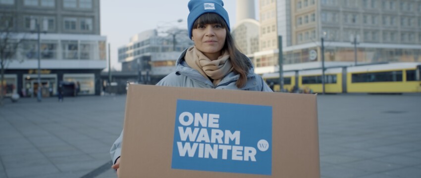 Eine dunkelhaarige Frau mit blauer Mütze schaut in die Kamera. Sie steht auf einem Platz und hat eine Winterjacke an. In der Hand hält sie einen Karton auf dem "One Warm Winter" steht.