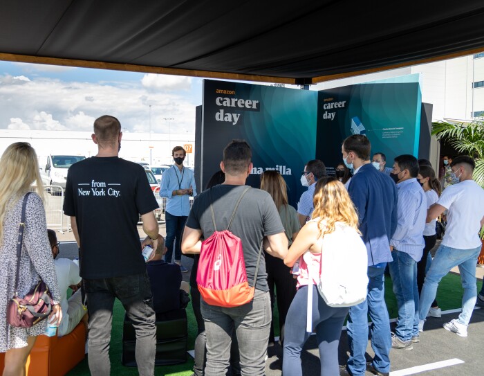 En España, el Career Day contó con una parte virtual y otra presencial, que se celebró en el nuevo centro logístico de Amazon en Illescas (Toledo). En primer plano un grupo de 20 personas de pie y se ve también que hay gente sentada en unos sillones de color naranja. Todas ellas de espaldas. Les habla una persona que está de pie y que se ve gesticular con mascarilla. De fondo el centro logístico de Amazon en Illescas. La gente está escuchando y en la parte derecha hay un cartel que llega al suelo y pone Amazon Career Day y Úlitma milla. Se ve el cielo azul con nubes blancas.