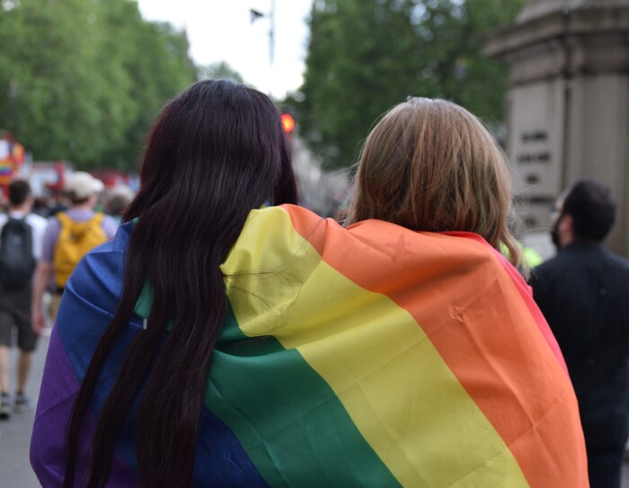 Es una foto con dos personas de pelo largo, moreno y rubio, de espaldas y tapados con una bandera del orgullo con el arcoiris. De fondo y desenfocados se ven personas andado en lo que parece un desfile. Se aprecia un autobús tojo de dos pisos con la bandera del orgullo. Por el autobús de dos pisos pensamos que es Londres.