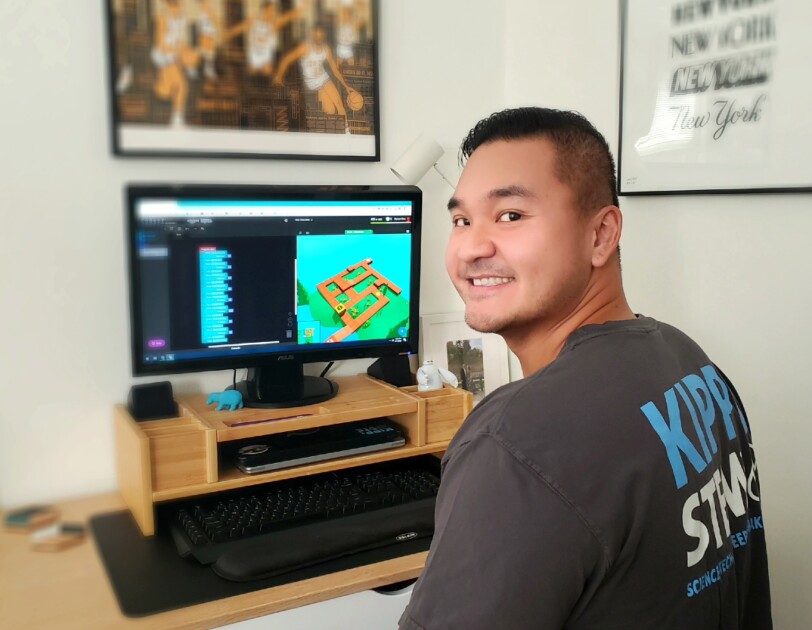 A man sitting at a desk with a computer on it turns around and smiles toward the camera.