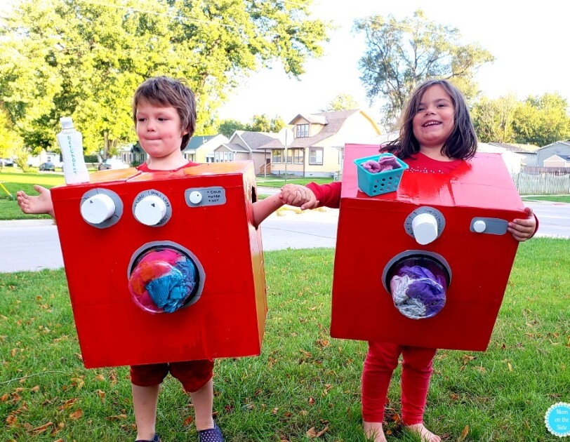 Two young children stand side-by-side, each wearing a red box. Both boxes have a clear "window" on the front that shows clothing, which look as though they're tumbling in the laundry. The child on the left has a bottle that says "detergent" on the top of the box, and two knobs. The child on the right has a small basket that looks like a laundry basket with socks, and one knob.