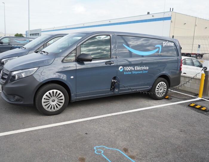 Una Mercedes Benz-Vans en un aparcamiento. La furgoneta es de color negro, con la sonrisa de color azul y la palabra Prime en blanco. Además, la furgoneta lleva la siguiente frase: 100% Eléctrico Envíos 'Shipment Zero'. La furgoneta está conectada a la red eléctrica y se está cargando la bateria.