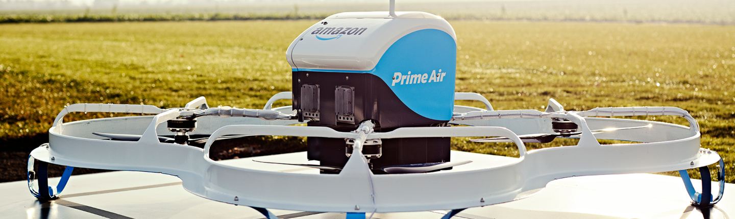 Biało-niebieski dron z logiem Prime Air oraz Amazon, umiejscowiony na metalowej powierzchni, w tle szerokie pole