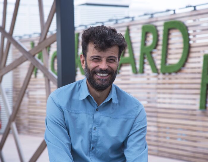 Alessandro Saccon, PR Manager di Amazon, sorride in primo sul terrazzo degli uffici Amazon di Milano. Sullo sfondo uno scorcio del terrazzo, con alcune lettere del motto 'Work hard' di Amazon.