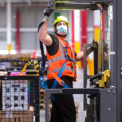 Dipendente di un magazzino Amazon. Indossa un casco, una mascherina chirurgica e un giubbino di sicurezza, si trova al centro di un macchinario. Alle sue spalle un cesto con dei pacchi Amazon.