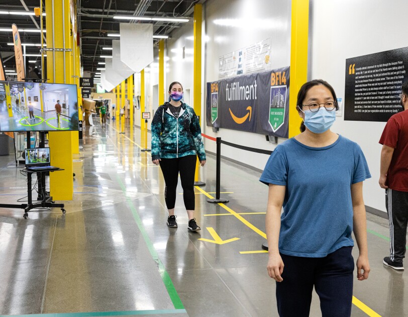 Amazon associates wearing face masks walk along a corridor of a fulfillment center. Behind them, a screen shows visual social distance assistance.