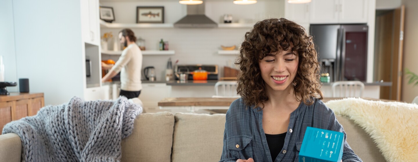 An Amazon customer smiles as they unbox a package on their couch.