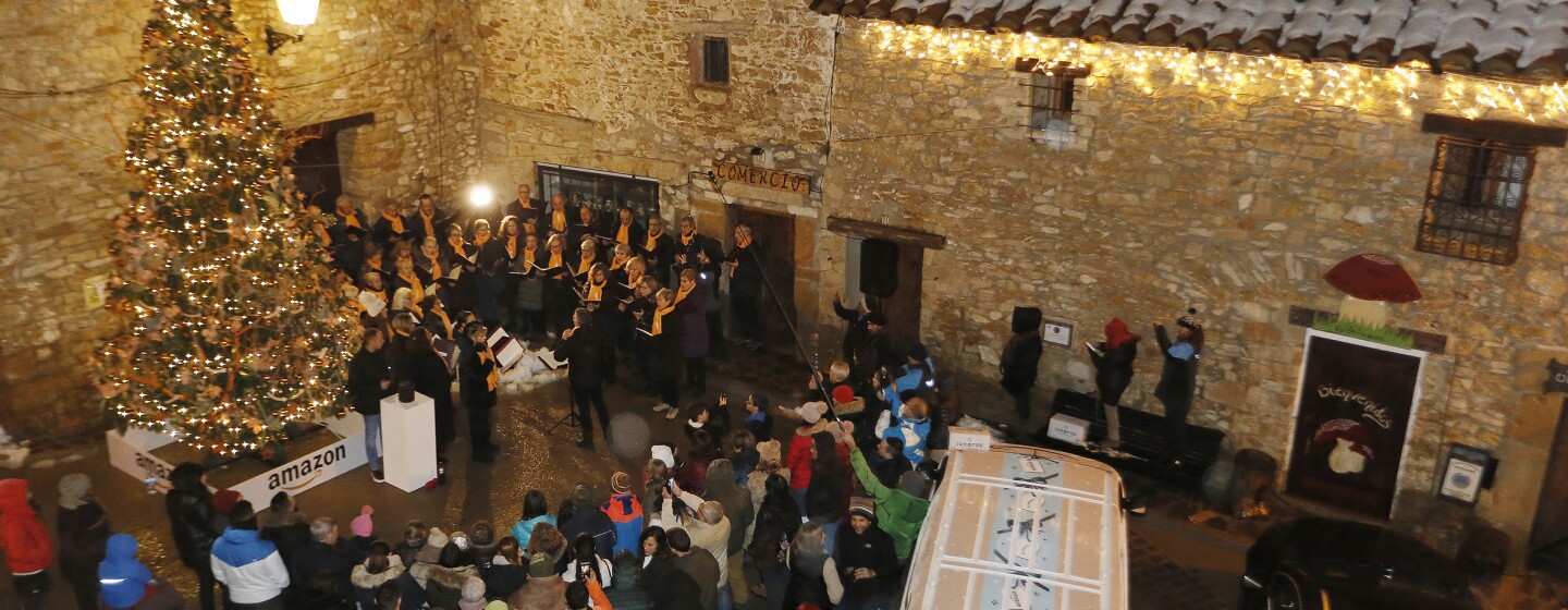En la plaza principal de Valdelinares rodeada de tres casa de piedra con nieve en el tejado se encuentra: un árbol de Navidad decorado con luces y detrás un coor de 40 cantantes vestidos de negro y con una bufanda de color amarillo. Rodeando el árbil y el coro, los vecinos del pueblo.