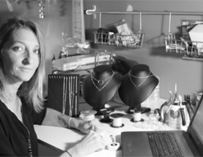 La fondatrice d'EmmaFashionNow se tient devant une table où se trouvent des bijoux finis ou en cours de création ainsi que son ordinateur. C'est une photo noir et blanc.