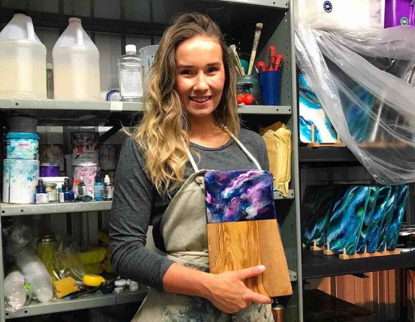 Kate Chesters holding one of her resin boards in her Liverpool-base studio, surrounding her are all of the utensils and products she uses to create her art.