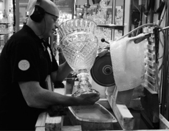 Un artisan est en train de polir un vase de cristal sur une machine électrique. Il porte un casque de protection sur les oreilles. Au fond on aperçoit des vitrines de créations en cristal. C'est une photo noir et blanc.