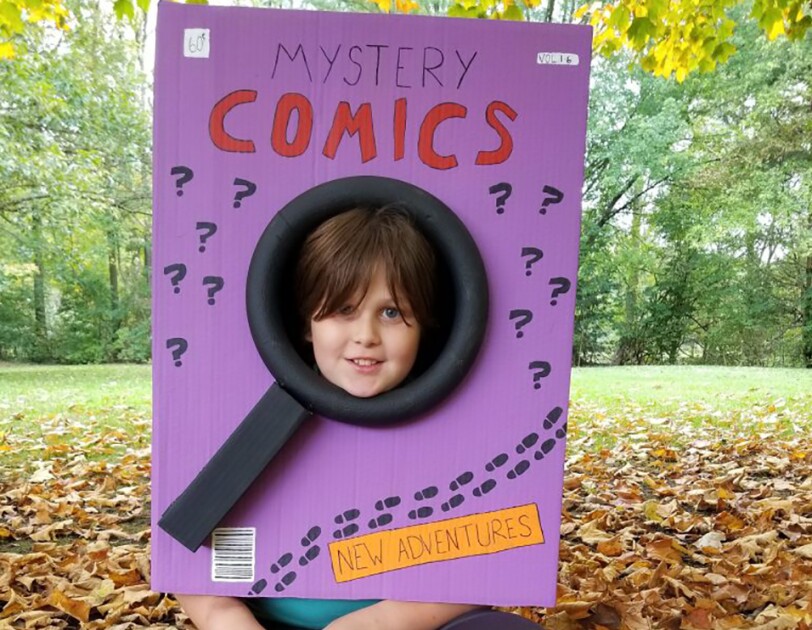 A comic book Halloween costume created from a box that was painted purple. It says "Mystery comics" and "new adventures," plus several question marks. The box is worn by a child, whose face peeks out from the center of a magnifying glass shape.